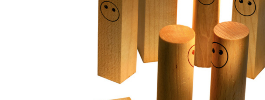 Holzklötze mit Gesichtern stehen vor einem weißen Hintergrund. Einer von ihnen liegt auf dem Boden.